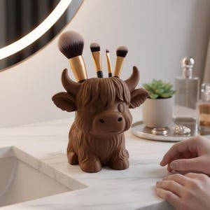 May include: A brown Highland cow-shaped makeup brush holder. The holder is made of a solid material and has a detailed design. Several makeup brushes with gold handles and black bristles are placed in the holder. The holder is on a white surface.