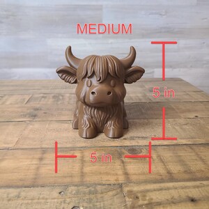 May include: A brown, medium-sized Highland cow figurine, approximately 5 inches tall and 5 inches wide. The figurine has detailed fur and horns, with the word "MEDIUM" above it. The cow is sitting on a wooden surface.