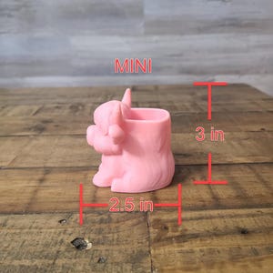 May include: A pink miniature Highland cow planter, 3 inches tall and 2.5 inches wide. The planter has a cylindrical opening for plants and the word "MINI" is printed on the side.