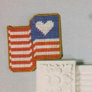 May include: Two small American flag-themed wall decorations with a heart in the center of each flag. The flags are red, white, and blue. The flags are hanging on a white wall with a decorative trim. There are red berries and green leaves in the background.