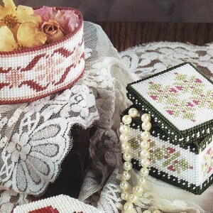 Peut inclure: Plusieurs boîtes en point de croix faites à la main. L'une est ovale et remplie de fleurs séchées, l'autre est carrée avec un couvercle. Les deux présentent des motifs complexes en rouge, blanc et vert. Un collier de perles est également visible.