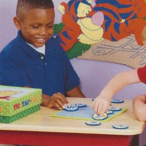 Könnte beinhalten: Zwei Kinder spielen Tic-Tac-Toe mit bunten Spielsteinen auf einem Holztisch. Die Spielbox ist grün und zeigt eine Comicfigur. Der Hintergrund zeigt eine bunte Wand mit einem Tigger-Wandbild.