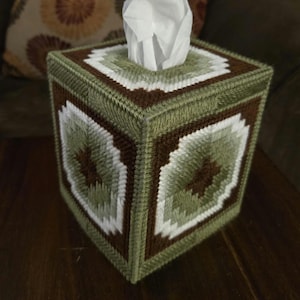 May include: A handmade tissue box cover in shades of green, brown, and white. The cube-shaped cover features a geometric design and a square opening at the top for tissue dispensing. The tissue box is sitting on a wooden surface.