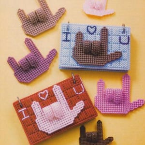 May include: Six colorful plastic canvas crafts featuring the American Sign Language symbol for "I love you". The crafts are in various colors, including pink, brown, and blue. The crafts are decorated with small hearts.
