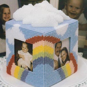 May include: A handmade, cube-shaped tissue box cover with a blue, white, red, and yellow stitched design. The cover features two photo frames with pictures of people. A white cloud-shaped embellishment sits on top.