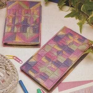 May include: Two needlepoint notebooks with geometric patterns in shades of pink, purple, yellow, and green. One notebook is open, and the other is closed. The notebooks are on a white surface.