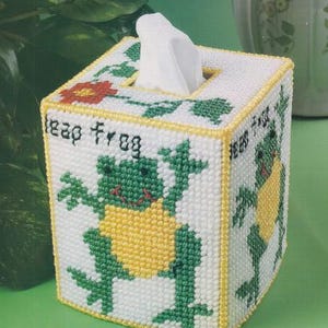 May include: White tissue box cover with a cross-stitch design featuring a green frog with a yellow belly. The words "Leap Frog" are stitched on the side. The top has a floral design and a yellow border.