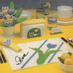 May include: A yellow tablecloth is set with a variety of items. A bouquet of fabric flowers, a napkin holder, and a placemat with a tulip design are visible. There are also glasses, plates, cutlery, and a bowl of lemons.