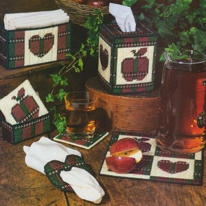 May include: A set of handmade craft items featuring a plaid and apple design. The set includes a tissue box cover, a napkin holder, coasters, and a napkin ring. The items are all made in a cream, green, red, and brown color scheme.