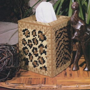 May include: A square tissue box cover with a woven design. The front features a leopard print pattern in black and brown against a beige background. The sides have a brown and black chevron pattern. A bundle of white tissues is visible.