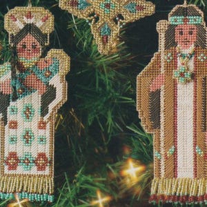 May include: Handmade Christmas ornaments featuring Native American figures. The ornaments are made with a needlepoint technique, showcasing detailed designs in various colors. The figures are adorned with beaded accents and fringe, adding a touch of elegance to the festive decor.