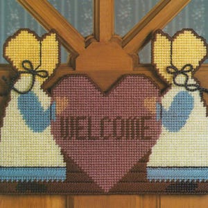 May include: Two figures holding a heart-shaped sign with the word "WELCOME" in brown letters. The figures are dressed in white dresses with blue accents and yellow bonnets.