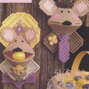 May include: Two handmade felt mouse ornaments with yellow, pink, purple and brown details. The mouse on the left is holding a yellow egg in its mouth. The mouse on the right is wearing a purple tie. Both mice are attached to a wooden surface.