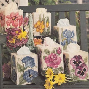 May include: Six handmade tissue box covers featuring floral embroidery designs. The covers showcase a variety of flowers, including roses, pansies, irises, and lilies. The covers are crafted from white fabric and have a delicate, vintage aesthetic.