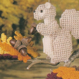 May include: A beige knitted squirrel holding acorns, displayed on a branch with autumn leaves in shades of yellow, orange, and brown. The background is a blurred autumnal scene. A charming, handcrafted decoration.