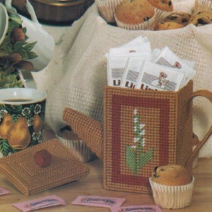 以下が含まれることがあります： 白い花柄の茶色のプラスチック製のキャンバス製のティーポット。ティーポットは砂糖の袋でいっぱいになっており、茶色の蓋付きの箱と小さなマフィンのあるテーブルの上に置かれています。