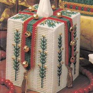 May include: A white plastic canvas tissue box cover, decorated for Christmas. It features green and red ribbon, gold bells, and green embroidered pine branches. A white tissue is visible at the top.