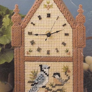 Puede incluir: Un reloj decorativo con un diseño cosido. El reloj presenta una combinación de colores beige y marrón, con números romanos que marcan las horas. La parte inferior del reloj muestra dos pájaros posados en una rama. El reloj tiene un techo triangular.