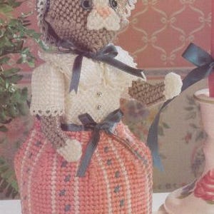 Puede incluir: Un calentador de té hecho a mano con forma de gato. El calentador es principalmente rosa con detalles blancos y azules. El gato lleva un vestido blanco con un lazo azul y un gorro. Diseñado para una tetera.