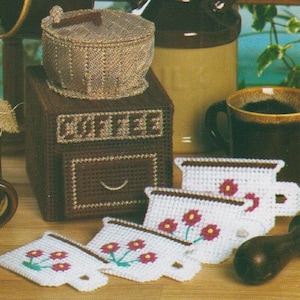May include: A set of four white ceramic coffee cup coasters with red flower designs. The coasters are shaped like coffee cups with handles. The coasters are displayed on a wooden surface next to a brown coffee can with a white label that says "Coffee".