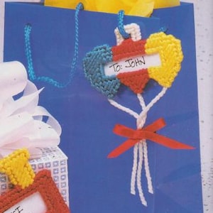 Puede incluir: Una bolsa de regalo azul con papel de seda amarillo y una etiqueta de regalo en forma de corazón hecha a mano que dice "To: John". La etiqueta está hecha de hilo azul, rojo y amarillo. Un lazo rojo está atado a la parte inferior de la etiqueta.