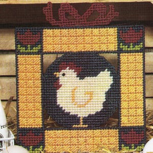 Op de afbeelding: Een ingelijst kruissteekpatroon met een witte kip met een rode kam en rode keellapjes, omringd door een zwarte achtergrond en een gele lijst met rode bloemen boven en onder.