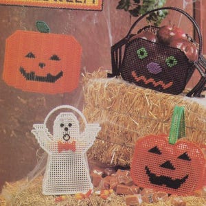 May include: A selection of Halloween plastic canvas decorations. Includes an orange pumpkin, a black spider basket filled with apples, a white ghost, and a pumpkin-shaped basket with a green handle. Sweets and wrapped candies are scattered around.