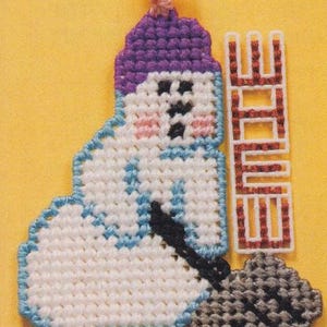May include: A handmade cross-stitch snowman with a purple hat and a black broom. The snowman has pink cheeks and a blue outline. Next to the snowman is a cross-stitch sign that says "WINTER".