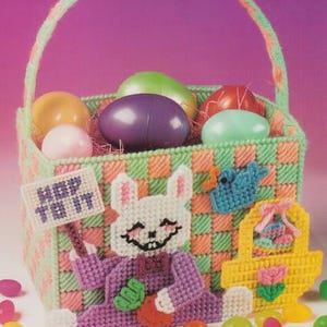 May include: A handmade Easter basket with a smiling bunny holding a sign that says "HOP TO IT." The basket is filled with colorful eggs and surrounded by jelly beans. The basket is made of plastic canvas in shades of green, orange, and pink.