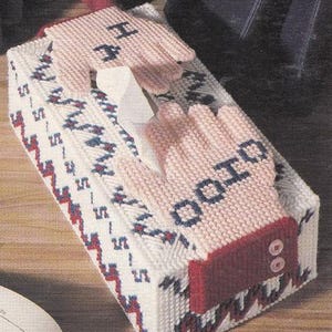 Op de afbeelding: Een witte tissueboxhoes met een geometrisch patroon in rood, wit en blauw. De hoes is versierd met twee roze gehaakte handschoenen met de woorden "Good" en "Hope" erop geborduurd.