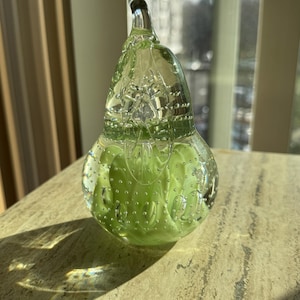 May include: A clear glass pear-shaped decorative object with a green interior. The glass has a bubbly texture and a long, curved stem. The object is sitting on a light-colored, textured surface.