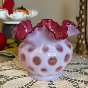 May include: A vintage pink glass vase with a ruffled rim and a polka dot pattern. The vase has a rounded body and a flared, ruffled top in a darker shade of pink. The vase is sitting on a white lace doily.