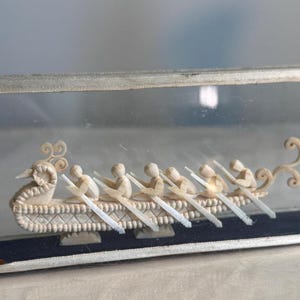 May include: A decorative ivory-colored boat with eight rowers, enclosed in a glass display case. The boat features intricate carvings and is displayed on a dark blue base. The case has a silver-colored frame.