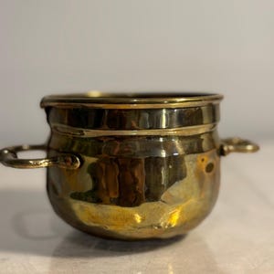 May include: A small, hammered brass pot with two loop handles. The pot has a rounded body and a slightly flared rim. The brass has a warm, golden color and a textured surface. The pot is likely vintage or antique.