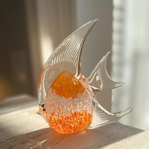 Peut inclure: Une sculpture de poisson en verre transparent avec des accents orange et blancs. Le poisson a de grandes nageoires transparentes et un corps texturé. La sculpture est exposée sur une surface claire.