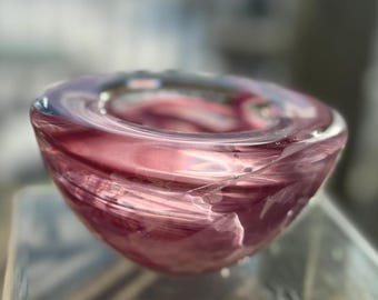May include: A handcrafted glass bowl with a rounded shape. The bowl features a blend of pink and purple hues, with a swirling pattern throughout the glass. The rim is a darker shade of purple. The bowl is likely used for decorative purposes.