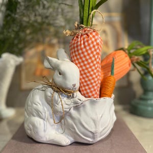 May include: A white ceramic bunny figurine holding a leaf-shaped bowl filled with decorative carrots. One carrot is orange and white gingham, and the others are orange. The bunny has a twine bow around its neck, and the carrots have green felt tops. Easter decor.