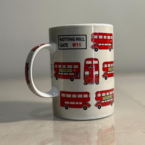 May include: White ceramic mug featuring red double-decker buses and the text "NOTTING HILL GATE W11". The handle also has a bus design. The mug is a souvenir or a themed item, likely for a London enthusiast.