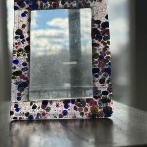 May include: A rectangular glass picture frame with a clear glass insert. The frame is decorated with colorful, speckled glass in shades of pink, blue, green, and purple. The frame is sitting on a dark surface.