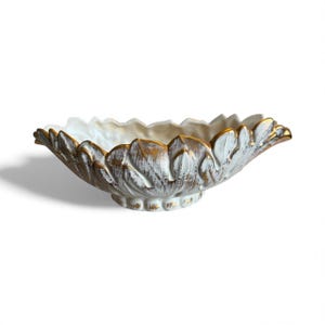 May include: An oval-shaped decorative bowl featuring a sculpted leaf pattern on the exterior. The surface has a distressed white finish with gold-toned accents highlighting the raised edges of the leaves and the scalloped rim of the vessel.