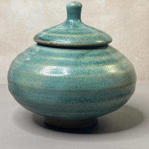 May include: A round, turquoise ceramic jar with a matching lid. The vessel features a glossy, mottled glaze with horizontal ridges along the body and a small, knob-like handle on top. The base is a contrasting dark, unglazed brown clay material.
