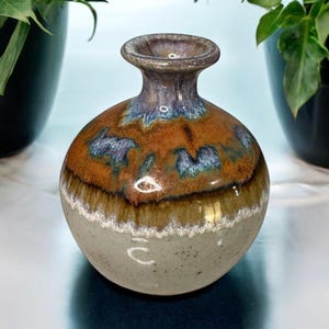 May include: A handcrafted ceramic vase with a round body and a narrow neck. The vase features a speckled gray base, transitioning to a brown and orange midsection with blue accents, and a dark purple rim. The vase is a decorative home accent.