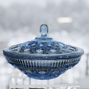 May include: A blue glass candy dish with a lid. The dish has a textured pattern of circles and diamonds. The lid has a small handle. The dish is sitting on a clear surface.