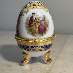 May include: A decorative egg-shaped trinket box with a hinged lid. The porcelain box features a painted scene of two people in period clothing. Gold accents and feet adorn the white and blue egg, adding to its ornate design.