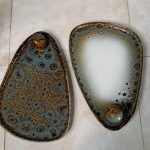 May include: Two ceramic serving trays with a speckled glaze. One tray is white with brown speckles and the other is brown with blue and green speckles. Both trays have a rounded, oval shape.