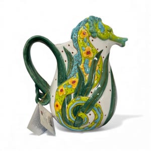 May include: A ceramic pitcher shaped like a seahorse with a white base, featuring hand-painted green seaweed, bright yellow flowers, and light blue accents. The handle is dark green, and a small white product tag hangs from the base of the handle.