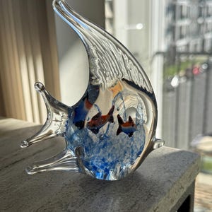 May include: A clear glass sculpture of a fish with a long, curved fin. The sculpture contains two small fish in shades of red and orange, set against a blue and white background. The fish is displayed on a gray surface.