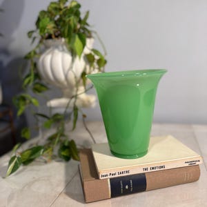 May include: A vibrant green glass vase with a flared top sits atop two vintage books. The books are stacked, with titles visible, and the vase is the focal point. A white decorative planter with greenery is in the background.