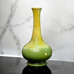 May include: A ceramic vase with a bulbous base and a slender, elongated neck. The surface features a smooth, glossy glaze that transitions from a deep, solid sage green at the bottom to a mottled, variegated yellow and gold pattern at the top.