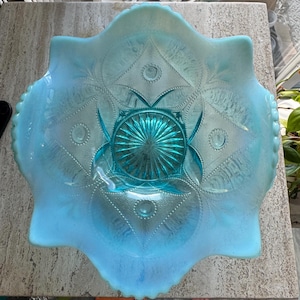 May include: A light blue, iridescent glass bowl with a starburst design in the center. The bowl has a scalloped edge and a detailed, embossed pattern. The bowl is approximately 10 inches in diameter.